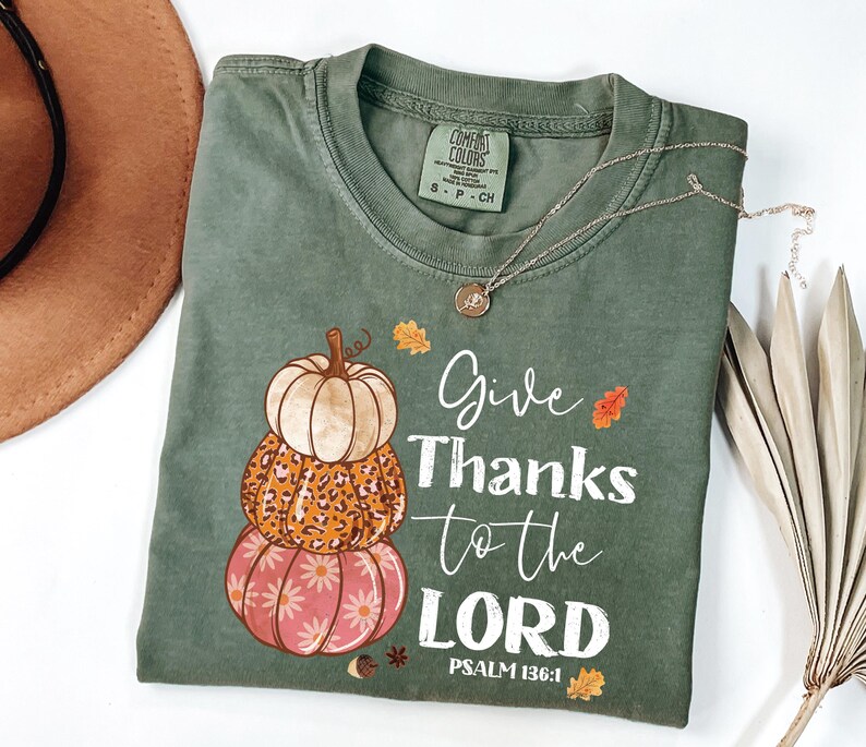 May include: Sage green t-shirt with a stacked pumpkin graphic in cream, leopard print, and pink. The shirt features the text "Give Thanks to the LORD" and "PSALM 136:1". A silver necklace with a pendant is draped across the shirt.