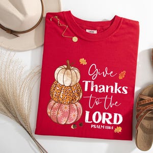 May include: A red t-shirt with a stacked pumpkin design and the text "Give Thanks to the LORD" in white. The pumpkins have different patterns and colors. The shirt is surrounded by a hat, sandals, and decorative elements.