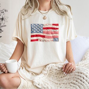 Peut inclure: T-shirt cr&egrave;me avec un drapeau am&eacute;ricain en patchwork sur le devant. Le drapeau comprend des rayures rouges et blanches et des &eacute;toiles bleues. Le t-shirt est port&eacute; de mani&egrave;re d&eacute;contract&eacute;e, sugg&eacute;rant le confort et un th&egrave;me patriotique.