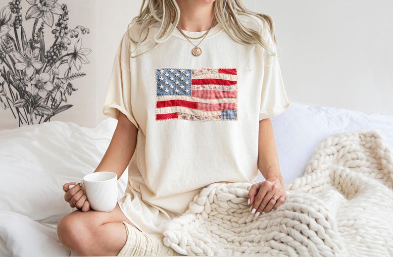 Peut inclure: T-shirt cr&egrave;me avec un drapeau am&eacute;ricain en patchwork sur le devant. Le drapeau comprend des rayures rouges et blanches et des &eacute;toiles bleues. Le t-shirt est port&eacute; de mani&egrave;re d&eacute;contract&eacute;e, sugg&eacute;rant le confort et un th&egrave;me patriotique.