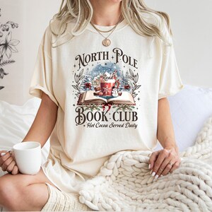 May include: Cream-colored t-shirt with a graphic that reads "North Pole Book Club" above an illustration of a book, a mug of hot cocoa, and a snowy scene. The text "Hot Cocoa Served Daily" is below. A white mug is held.