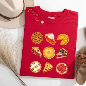 May include: A red t-shirt featuring watercolor illustrations of various pies, including apple, pumpkin, and cherry. The shirt is accompanied by a beige hat, gold necklace, and brown sandals. A sprig of dried grass is also present.
