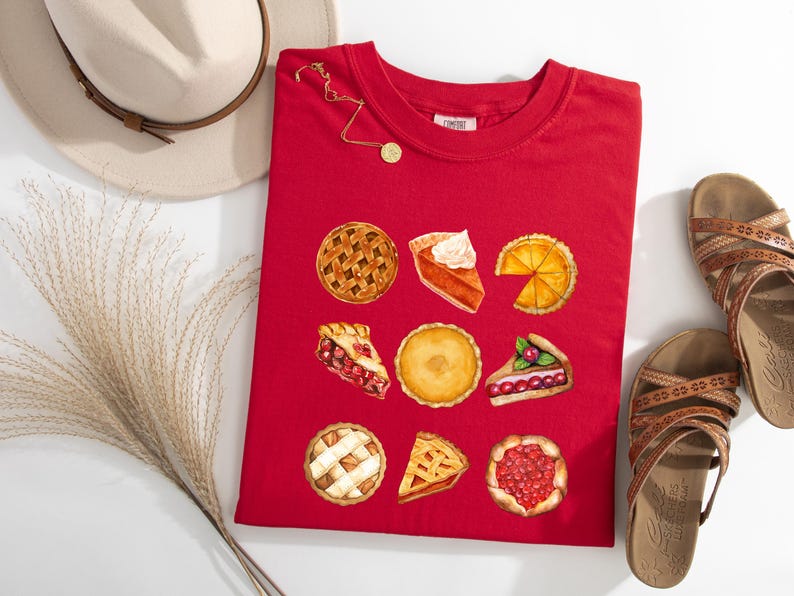 May include: A red t-shirt featuring watercolor illustrations of various pies, including apple, pumpkin, and cherry. The shirt is accompanied by a beige hat, gold necklace, and brown sandals. A sprig of dried grass is also present.