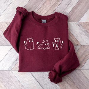 May include: A maroon sweatshirt featuring a white graphic of three cats dressed as ghosts. The cats are in different poses, with one sitting, one standing, and one with its arms up. The sweatshirt has a crew neck and long sleeves.