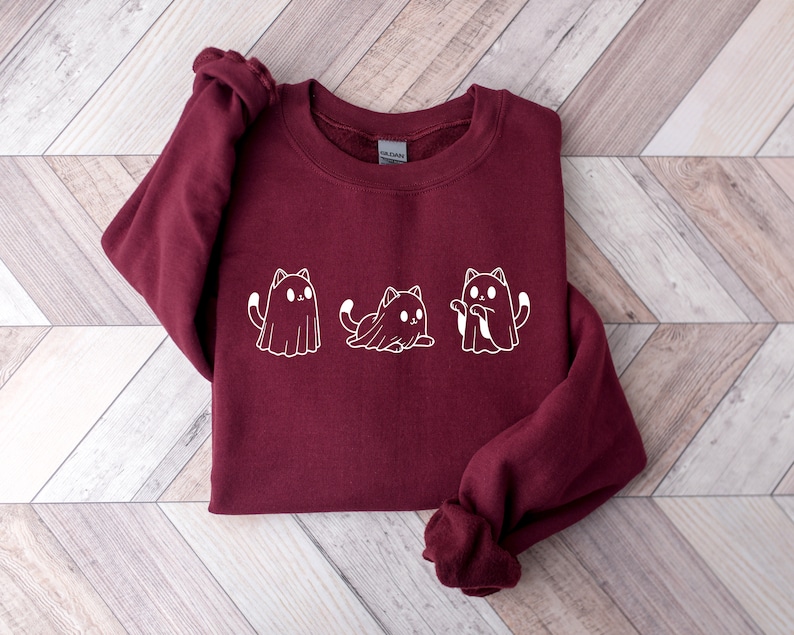 May include: A maroon sweatshirt featuring a white graphic of three cats dressed as ghosts. The cats are in different poses, with one sitting, one standing, and one with its arms up. The sweatshirt has a crew neck and long sleeves.