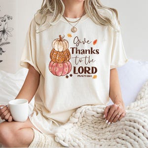 May include: Cream-colored t-shirt featuring a stacked pumpkin design in shades of orange and pink. The shirt has the text "Give Thanks to the LORD" with "Psalm 118:1" below. The shirt is worn by a person holding a white mug.