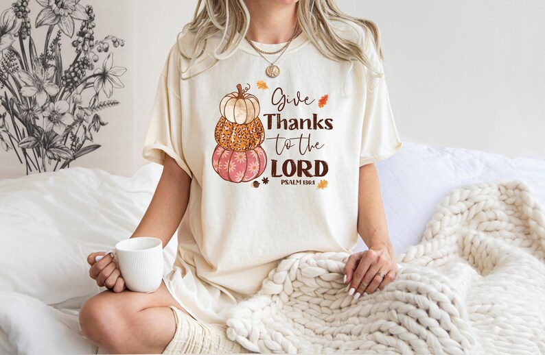 May include: Cream-colored t-shirt featuring a stacked pumpkin design in shades of orange and pink. The shirt has the text "Give Thanks to the LORD" with "Psalm 118:1" below. The shirt is worn by a person holding a white mug.