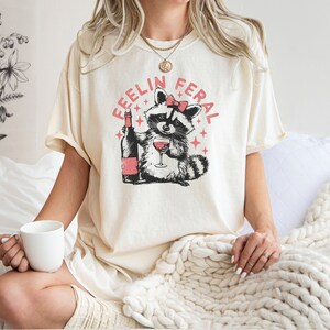 K&ouml;nnte beinhalten: Ein wei&szlig;es T-Shirt mit einem Aufdruck eines Waschb&auml;ren, der eine rosafarbene Schleife tr&auml;gt und ein Glas Wein h&auml;lt. Der Text "Feelin Feral" ist in Rosa um den Waschb&auml;ren herum geschrieben.