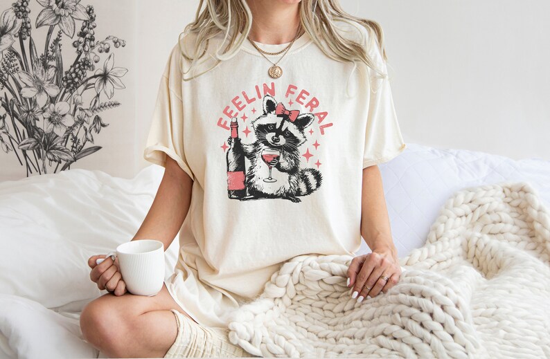 K&ouml;nnte beinhalten: Ein wei&szlig;es T-Shirt mit einem Aufdruck eines Waschb&auml;ren, der eine rosafarbene Schleife tr&auml;gt und ein Glas Wein h&auml;lt. Der Text "Feelin Feral" ist in Rosa um den Waschb&auml;ren herum geschrieben.