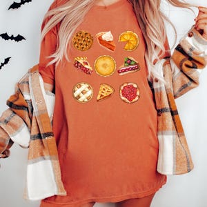 May include: Burnt orange t-shirt featuring a collection of pie illustrations. The shirt is paired with a plaid flannel shirt. Black bat cutouts are on the wall behind the shirt.
