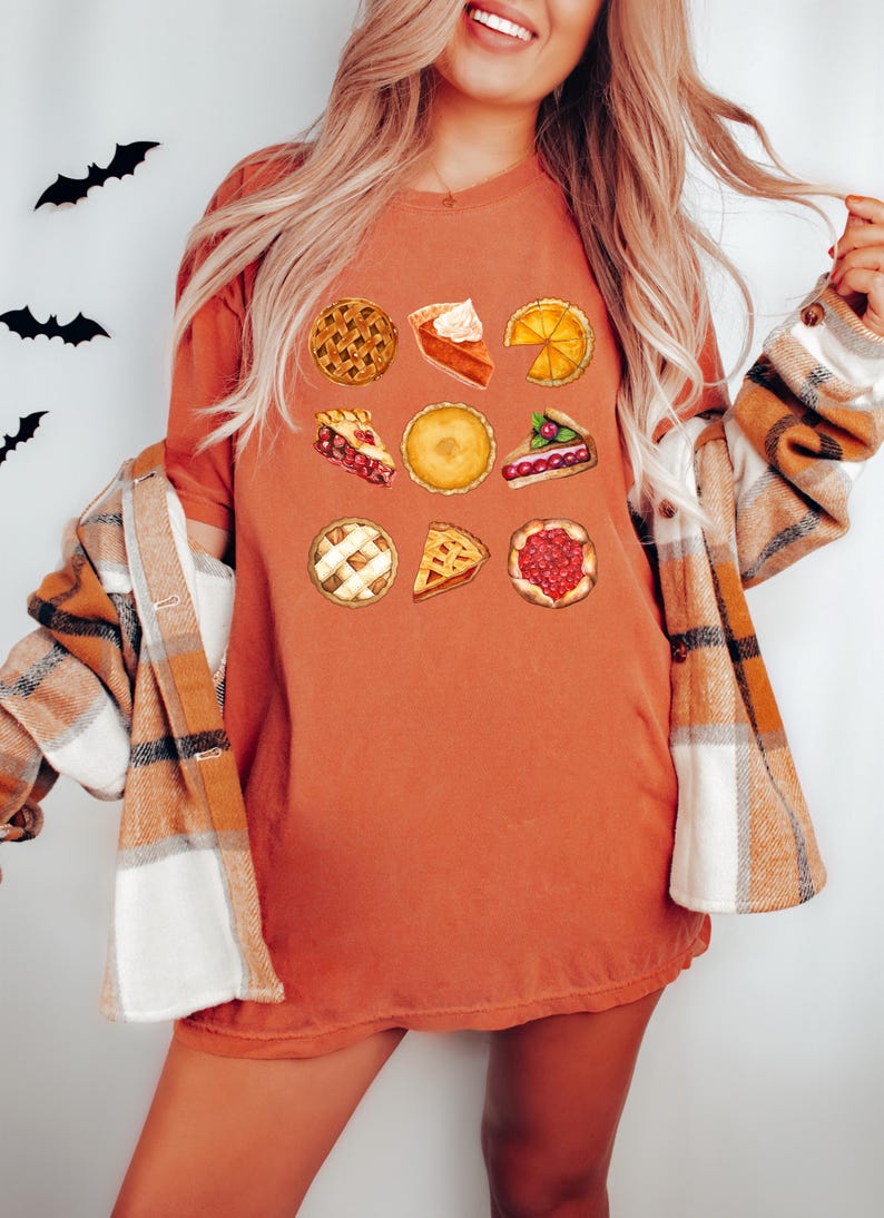 May include: Burnt orange t-shirt featuring a collection of pie illustrations. The shirt is paired with a plaid flannel shirt. Black bat cutouts are on the wall behind the shirt.
