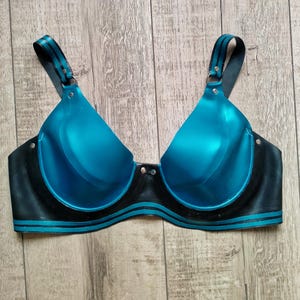 Puede incluir: Un sujetador teal y negro con un acabado brillante. El sujetador presenta copas moldeadas, tirantes ajustables y una banda bajo el busto negra con rayas teal. Los tirantes y la banda bajo el busto tienen un diseño a rayas. El sujetador está hecho de un material brillante.