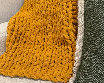 May include: A mustard yellow chunky knit throw blanket with a white fringe on a light gray couch. The blanket is next to a dark green pillow with a herringbone pattern.