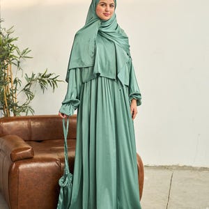 May include: A woman wearing a sage green satin dress and matching head covering. The long-sleeved dress has a flowing skirt and a small matching handbag. The outfit is a modest style, suitable for special occasions or everyday wear.