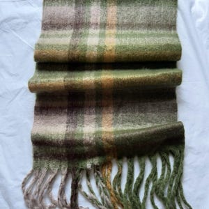 Op de afbeelding: Een groene, bruine en beige geruite sjaal met franjes. De sjaal is opgevouwen en ligt op een witte achtergrond. Het kleurenpalet omvat verschillende tinten groen, bruin, beige en een vleugje geel.