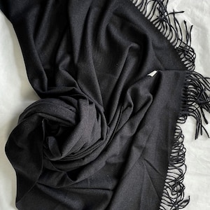 May include: A black fringed scarf made of a soft, warm fabric. The scarf is folded and draped on a white surface.