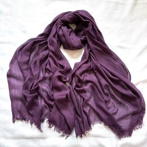 May include: A deep purple scarf made of a lightweight, semi-sheer fabric. The scarf features a soft, crinkled texture throughout its length and is finished with delicate, frayed fringe edges along the bottom hem, creating a casual appearance.