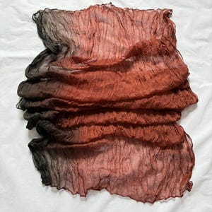 May include: A folded, textured scarf with a color gradient from black to reddish-brown. The fabric appears lightweight and has a crinkled texture, creating a layered effect. The scarf is displayed on a white surface.