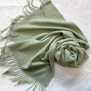 May include: A soft, sage green scarf with fringed edges. The scarf is folded and draped on a white surface.
