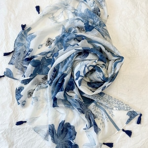 Könnte beinhalten: Weißer Schal mit blauem Blumenmuster und marinefarbenen Quasten. Der Schal hat ein zartes Design aus blauen Blumen und Blättern, das eine weiche und luftige Ästhetik erzeugt. Die Kanten sind mit kleinen marinefarbenen Quasten versehen.