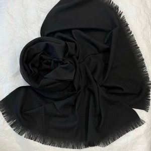 May include: A black fringed scarf made of a soft, lightweight fabric. The scarf is folded and draped in a way that shows the texture of the fabric and the fringe detail.