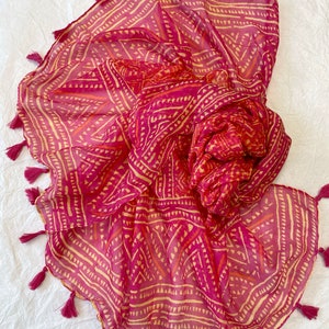 以下が含まれることがあります： フリンジが付いたピンクと黄色の模様の入ったスカーフ。スカーフは軽量の生地で作られており、幾何学的なデザインが施されています。