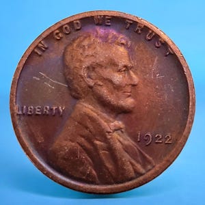 Puede incluir: Primer plano de un centavo Lincoln de 1922 de color cobre. La moneda presenta un perfil de Abraham Lincoln, la palabra "LIBERTY" y la inscripción "IN GOD WE TRUST". El año "1922" es visible.