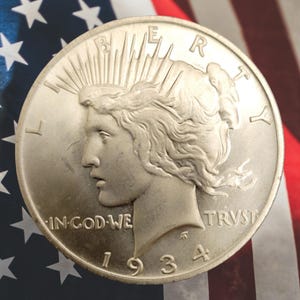 May include: A silver Peace dollar coin from 1934, featuring a profile of Lady Liberty and the inscription "IN GOD WE TRUST." The coin is set against a backdrop of the American flag, with its stars and stripes.