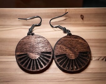 Puede incluir: Un par de pendientes de madera con una mancha marrón oscura. Los pendientes son redondos y tienen un diseño geométrico tallado en la madera. Los pendientes están unidos a ganchos de plata.