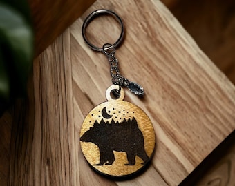 May include: A wooden keychain with a bear silhouette, a crescent moon, and stars against a gold background. The keychain has a silver ring and a small silver leaf charm.