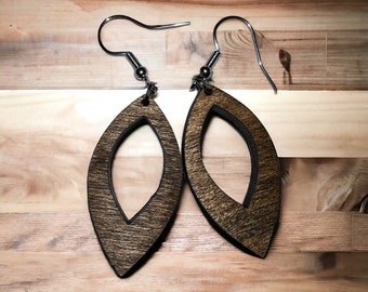 Puede incluir: Un par de pendientes de madera con forma de hoja y un diseño recortado. Los pendientes son marrones con un contorno marrón oscuro y tienen ganchos plateados.