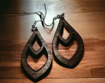 Puede incluir: Un par de pendientes de madera con forma de lágrima y un diseño recortado. Los pendientes son de color marrón oscuro y tienen ganchos plateados.