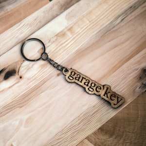 Könnte beinhalten: Ein Holzschlüsselanhänger mit der Aufschrift "garage key". Der Schlüsselanhänger ist an einem silbernen Metallring befestigt.