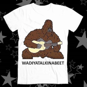 Peut inclure: T-shirt blanc avec une image de dessin animé d'un sasquatch brun jouant de la guitare. Le texte "WADIYATALKINABEET" est imprimé sous l'image.