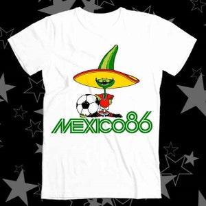 Puede incluir: Camiseta blanca con un personaje de dibujos animados verde que lleva un sombrero y sostiene un balón de fútbol. El texto "MEXICO 86" está impreso en verde debajo del personaje.