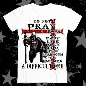 May include: White t-shirt featuring a red cross and a knight in armour. The text reads "Do not pray for an easy life pray for the strength to endure a difficult one."