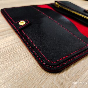 Op de afbeelding: Zwarte leren portemonnee met rode stiksels en een gouden rits. De portemonnee heeft een klep sluiting en een rood interieur.