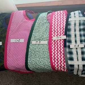 May include: Five different fabric bibs with different patterns and colors. The bibs are labeled with numbers 0-1, 0-2, 0-3, 0-4, and 0-5.