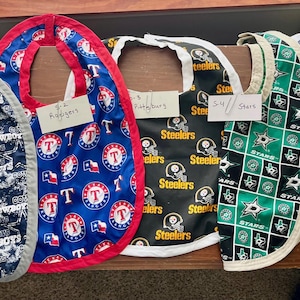 Peut inclure: Une collection de bavoirs pour bébés avec différents logos et motifs d'équipes sportives. Les bavoirs incluent les Cowboys de Dallas, les Rangers du Texas, les Steelers de Pittsburgh, les Stars de Dallas et les Astros de Houston.