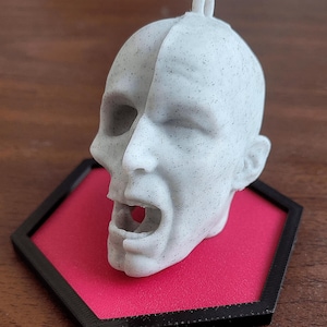 Op de afbeelding: Een lichtgrijze, 3D-geprinte sculptuur van een schreeuwend menselijk hoofd. Het hoofd is gemonteerd op een rode, zeshoekige basis met een zwarte rand. De sculptuur heeft een lus aan de bovenkant.