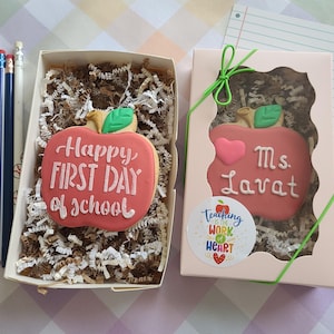 May include: Two apple-shaped sugar cookies in boxes. One cookie says "Happy FIRST DAY of school" in white icing. The other says "Ms. Lavat" with a pink heart. Both have green leaves. A sticker reads "Teaching is the WORK of HEART". Three pencils are included.