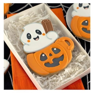 May include: A sugar cookie shaped like a pumpkin mug filled with white icing and a brown cinnamon stick. The pumpkin is orange with a dark gray face, and the white icing features a smiling ghost-like face with black eyes and orange cheeks.