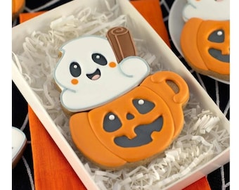 May include: A sugar cookie shaped like a pumpkin mug filled with white icing and a brown cinnamon stick. The pumpkin is orange with a dark gray face, and the white icing features a smiling ghost-like face with black eyes and orange cheeks.