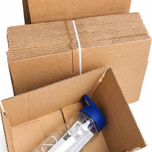 May include: A clear plastic water bottle with a blue cap and a white label inside a cardboard box. Several stacks of folded cardboard boxes are in the background, secured with a white strap. The bottle has a logo.