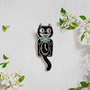 May include: Black enamel pin of a cat with a clock face on its belly, wearing a teal bow tie. The cat's eyes are white with black pupils, and it has a small pink heart on its chest.