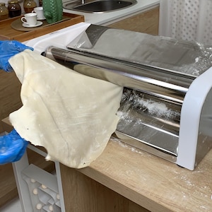 Può includere: Una macchina per la pasta in acciaio inossidabile con un telaio bianco è mostrata, con un foglio di pasta che viene fatto passare attraverso i rulli. La macchina è appoggiata su un piano di lavoro in legno, con una spatola bianca nelle vicinanze. La scena suggerisce la preparazione di pasta fatta in casa.