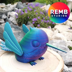May include: A 3D printed hummingbird figurine with iridescent blue, green, and purple colors. The figurine has a long, thin beak and is perched on a wooden base.