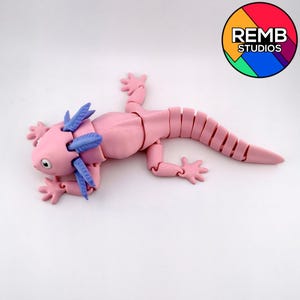 May include: A pink articulated axolotl toy with blue gills and small, detailed feet. The toy is lying on a white surface. The axolotl has a segmented body and a smiling face. The REMB STUDIOS logo is in the upper right corner.