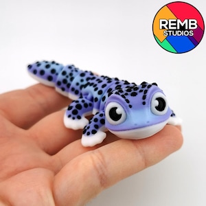 Puede incluir: Figurita de gecko artesanal, en tonos morados y azules, salpicada de puntos negros. El gecko tiene ojos grandes y expresivos y detalles blancos en las patas. El logotipo de REMB STUDIOS está en la esquina superior derecha.