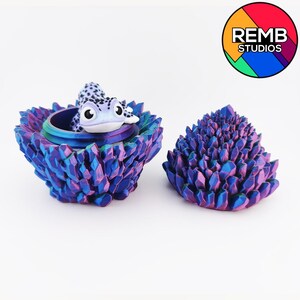 May include: A decorative, iridescent blue and purple container with a matching lid. A small, spotted frog figurine sits inside the container. The container and lid have a crystal-like texture. The logo for REMB STUDIOS is in the upper right corner.